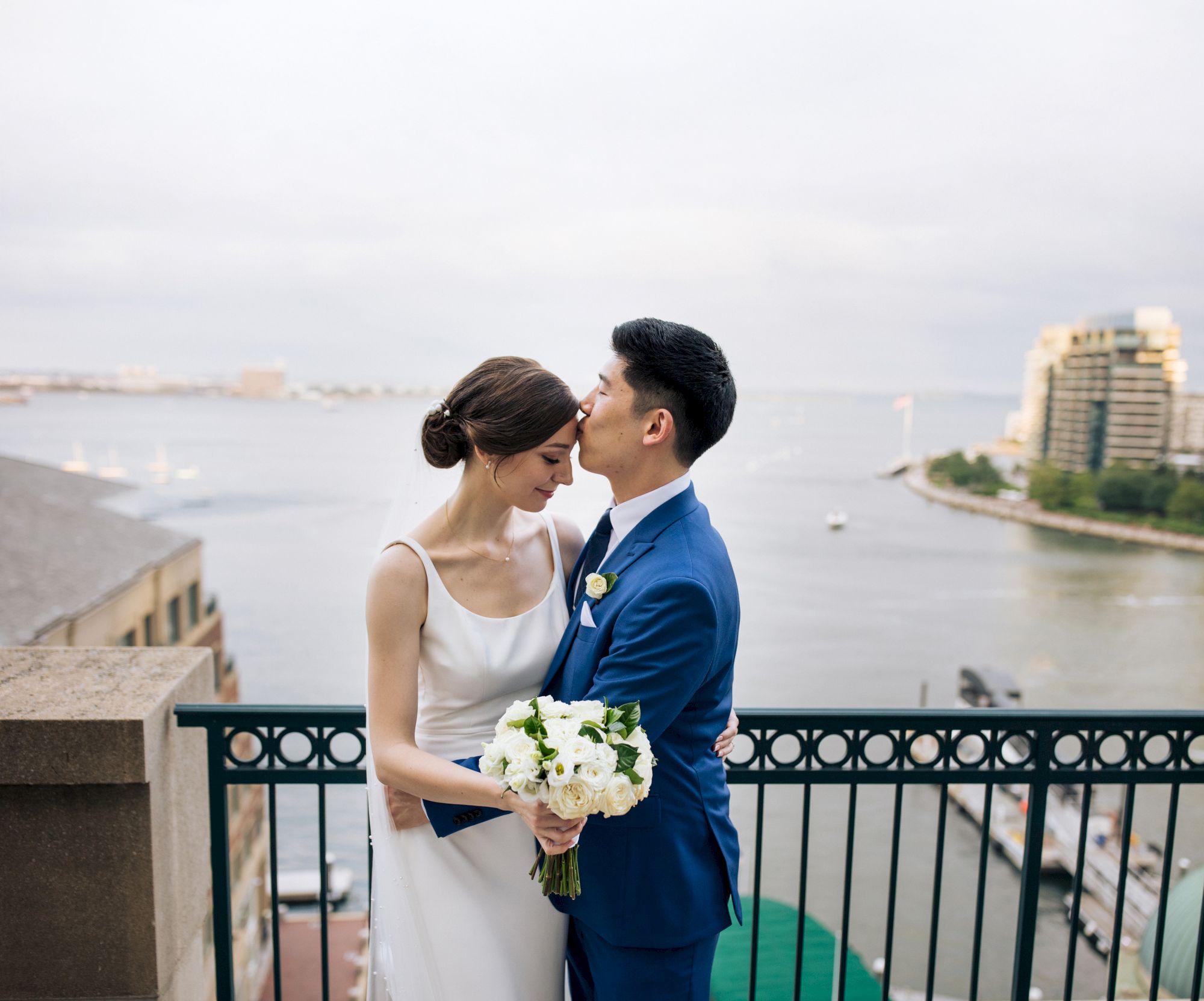 A couple in wedding attire embrace on a balcony overlooking a body of water and cityscape, with the groom kissing the bride's forehead.