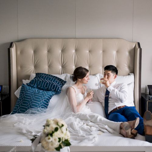 A couple in wedding attire is relaxing on a bed with drinks, surrounded by elegant decor and flowers, creating a celebratory atmosphere.