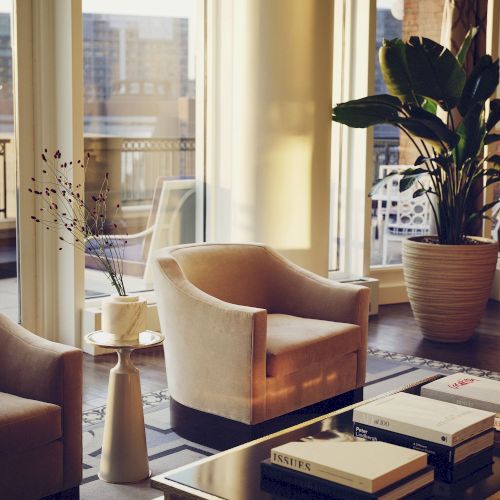 A stylish living room features two beige chairs, a coffee table with books, a large potted plant, and a window view of a cityscape.