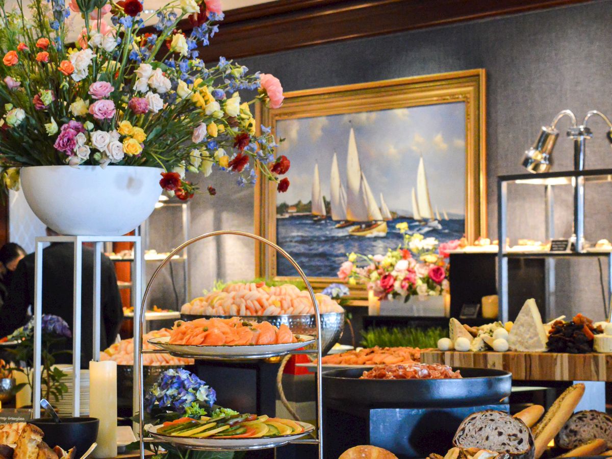 A buffet spread with a variety of foods, elegant flower arrangements, and a painting of sailboats on the wall.