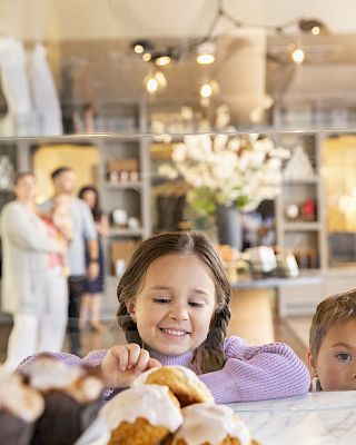 A child joyfully looks at pastries with icing, while another child peers over in a well-lit bakery setting.