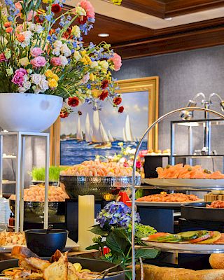 A vibrant buffet setup with flowers, seafood, and various dishes, set against a nautical-themed painting in a luxurious setting.